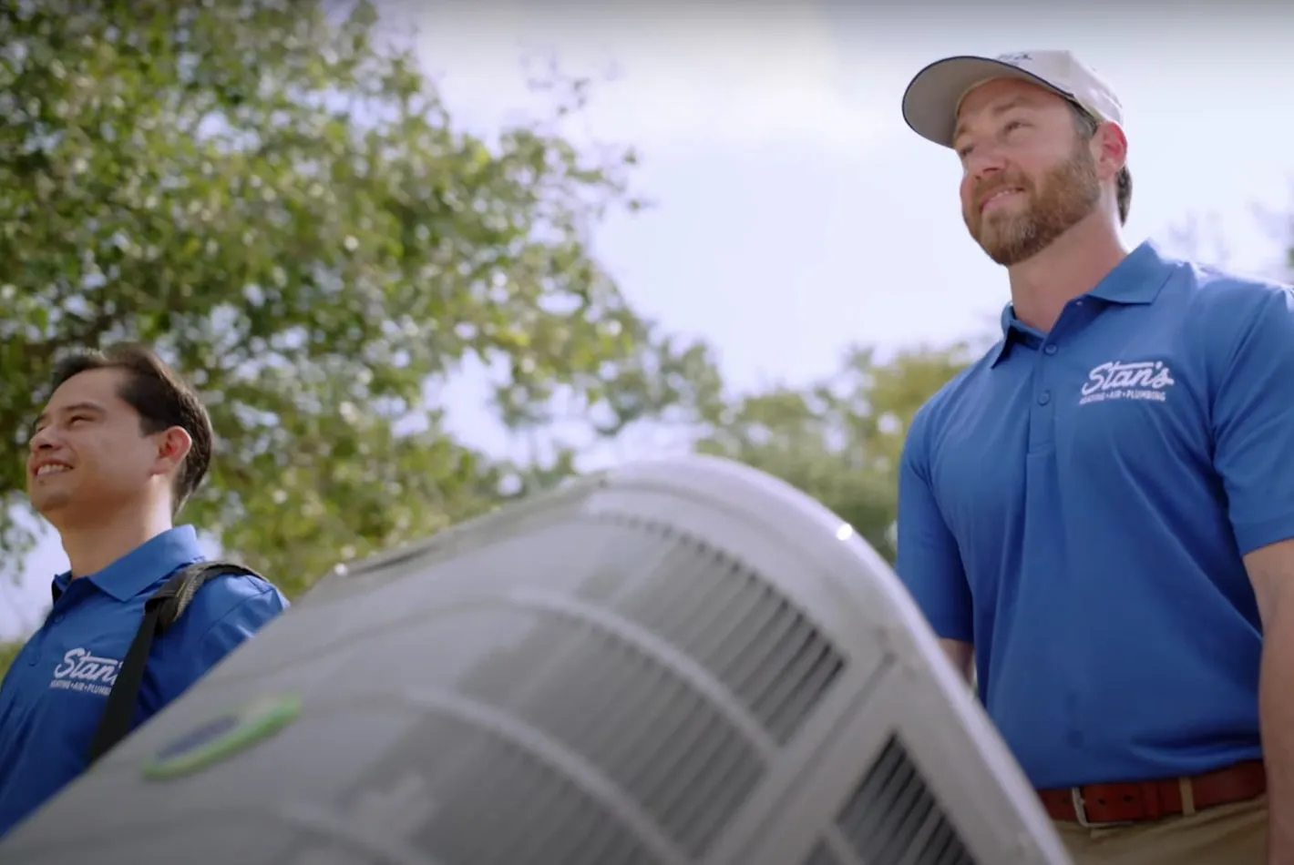 Two male technicians from Stan's are standing outdoors next to an air conditioning unit. Both are wearing blue Stan's polo shirts and looking ahead with smiles. The background features trees and a bright sky, creating a pleasant atmosphere.