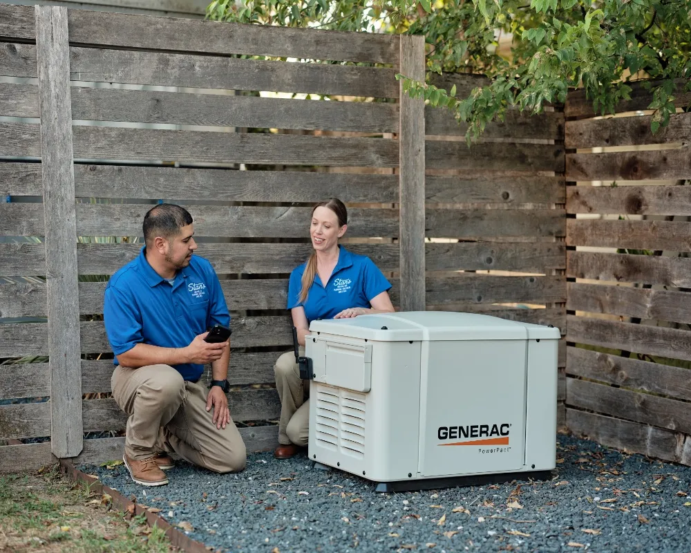 Two technicians from Stan's, one male and one female, are kneeling on gravel next to a Generac generator outside a house. The male technician is holding a smartphone, and they appear to be discussing something related to the generator.