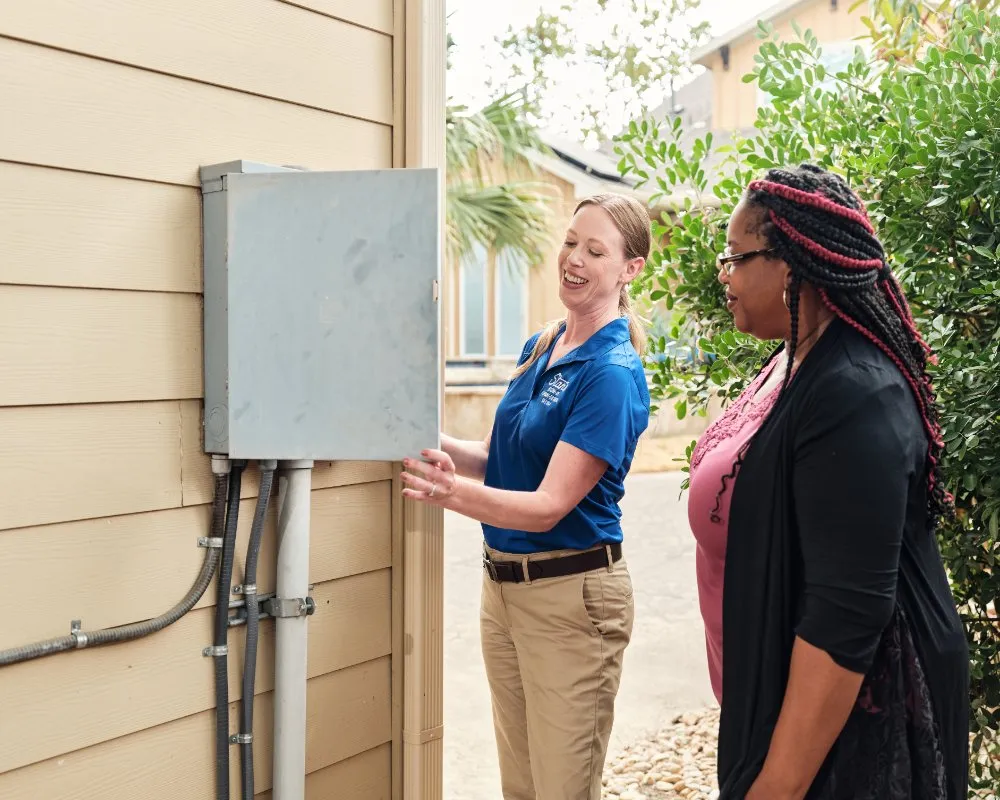 "A Stan's Heating, Plumbing, and Electrical technician in a blue company polo shirt and khaki pants is showing an outdoor electrical panel to a customer. The technician and the customer are smiling, and the panel is attached to the side of a beige house."