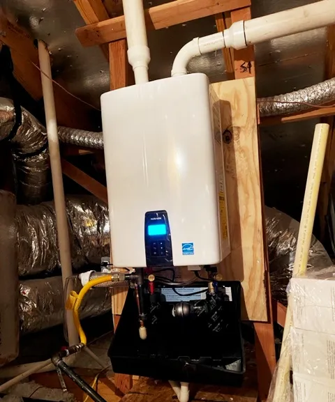 Tankless water heater mounted on a wall in an attic with exposed pipes and insulation.