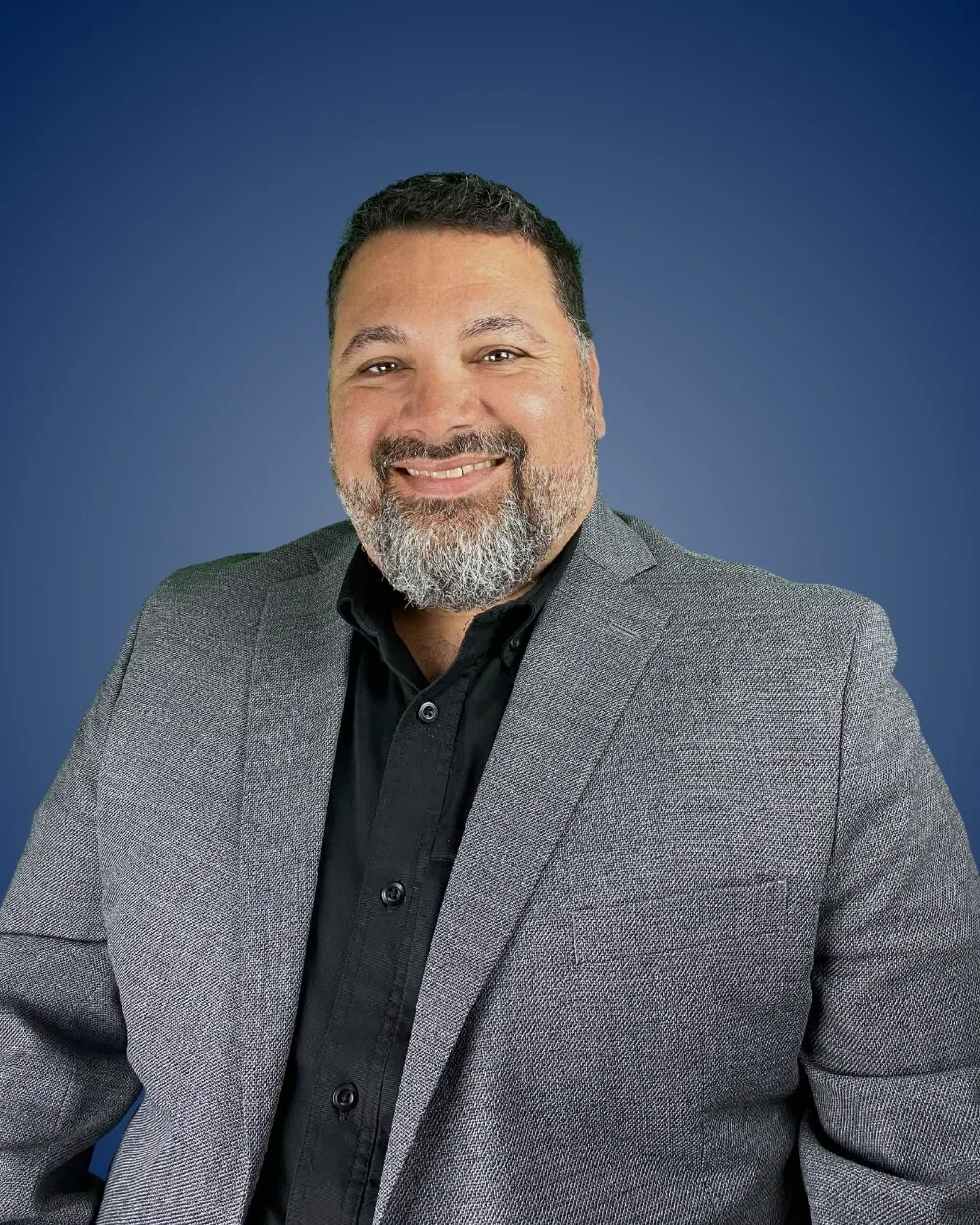 Roland Arrisola in gray blazer with a beard smiling against blue background.