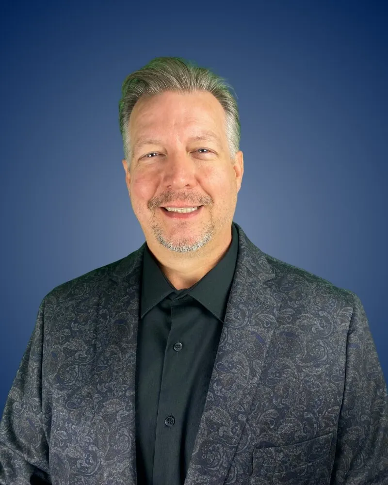 Brian Cardwell in paisley jacket smiling against blue background.