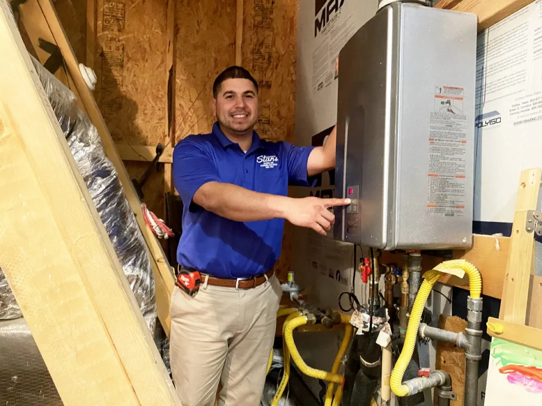"A Stan's Heating, Plumbing, and Electrical technician wearing a blue company polo shirt and khaki pants is standing in an attic, pointing at a tankless water heater. The technician is smiling and holding a tool belt."