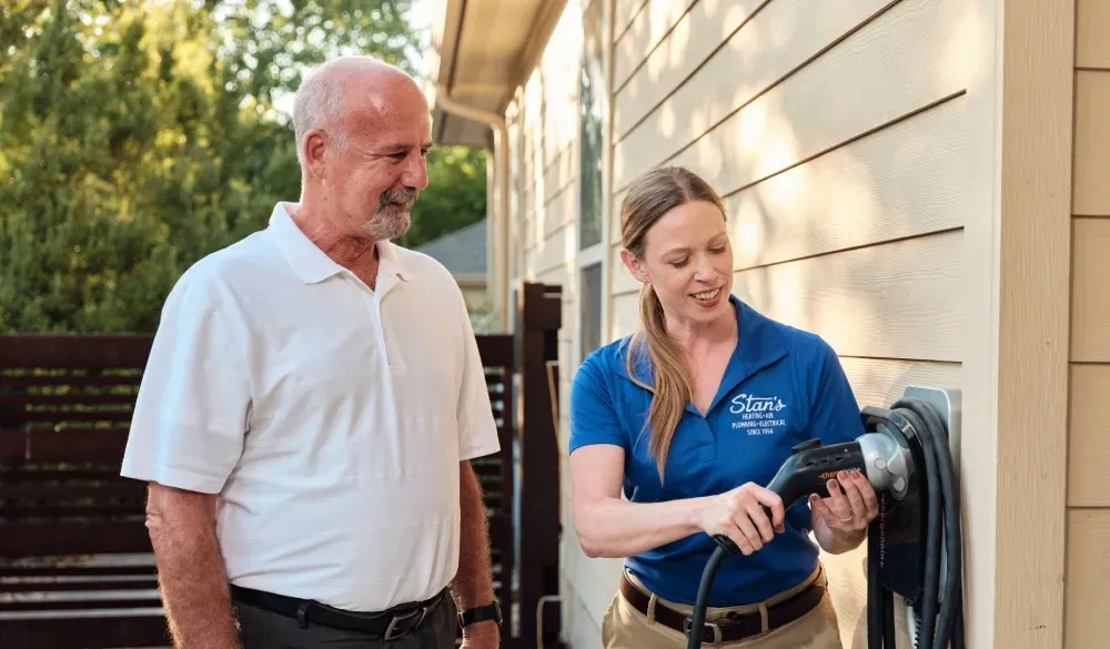 A woman in a blue shirt demonstrates a wall-mounted device to an older man outdoors.