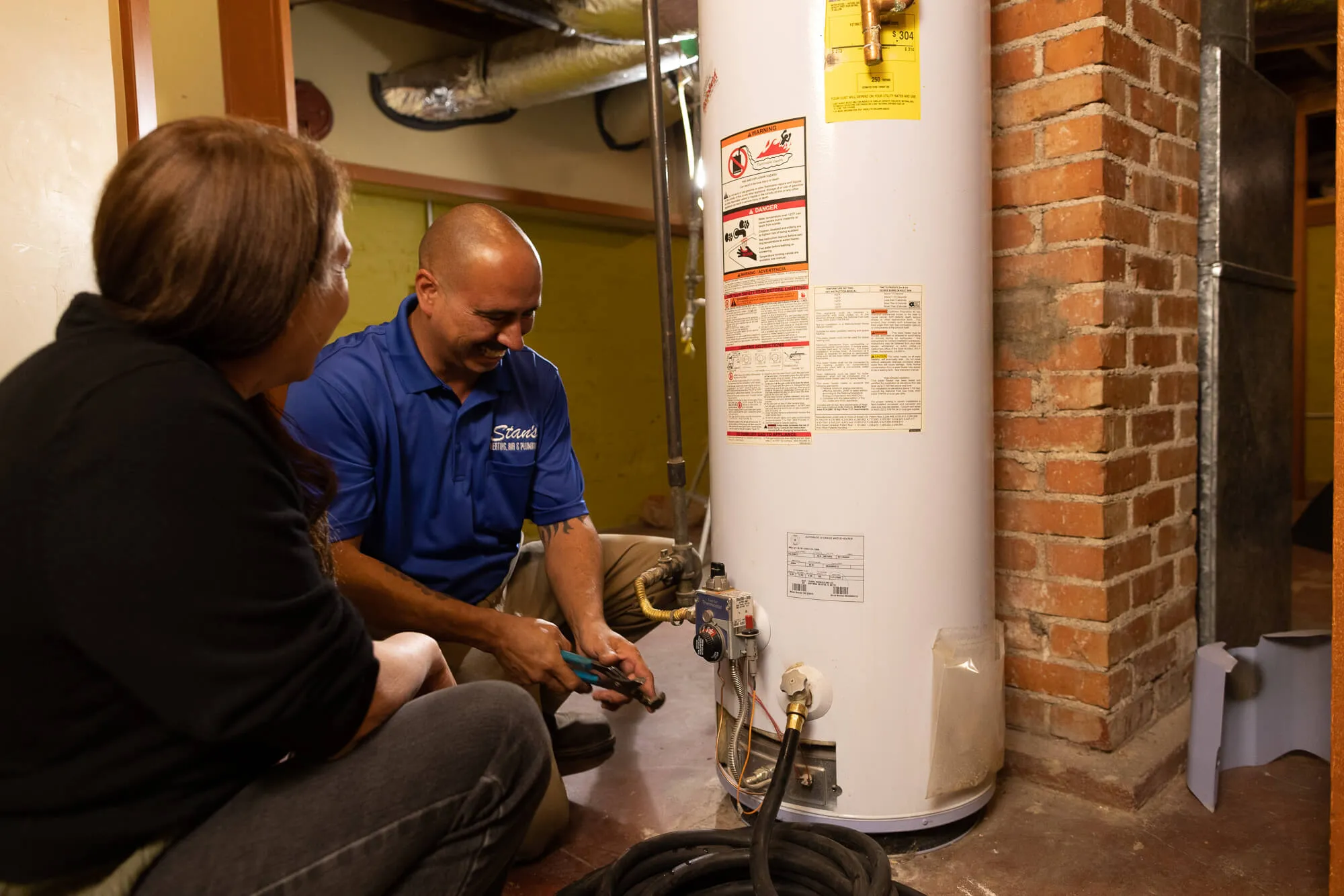 A technician in a blue shirt is inspecting a water heater while talking to a seated person in a basement.