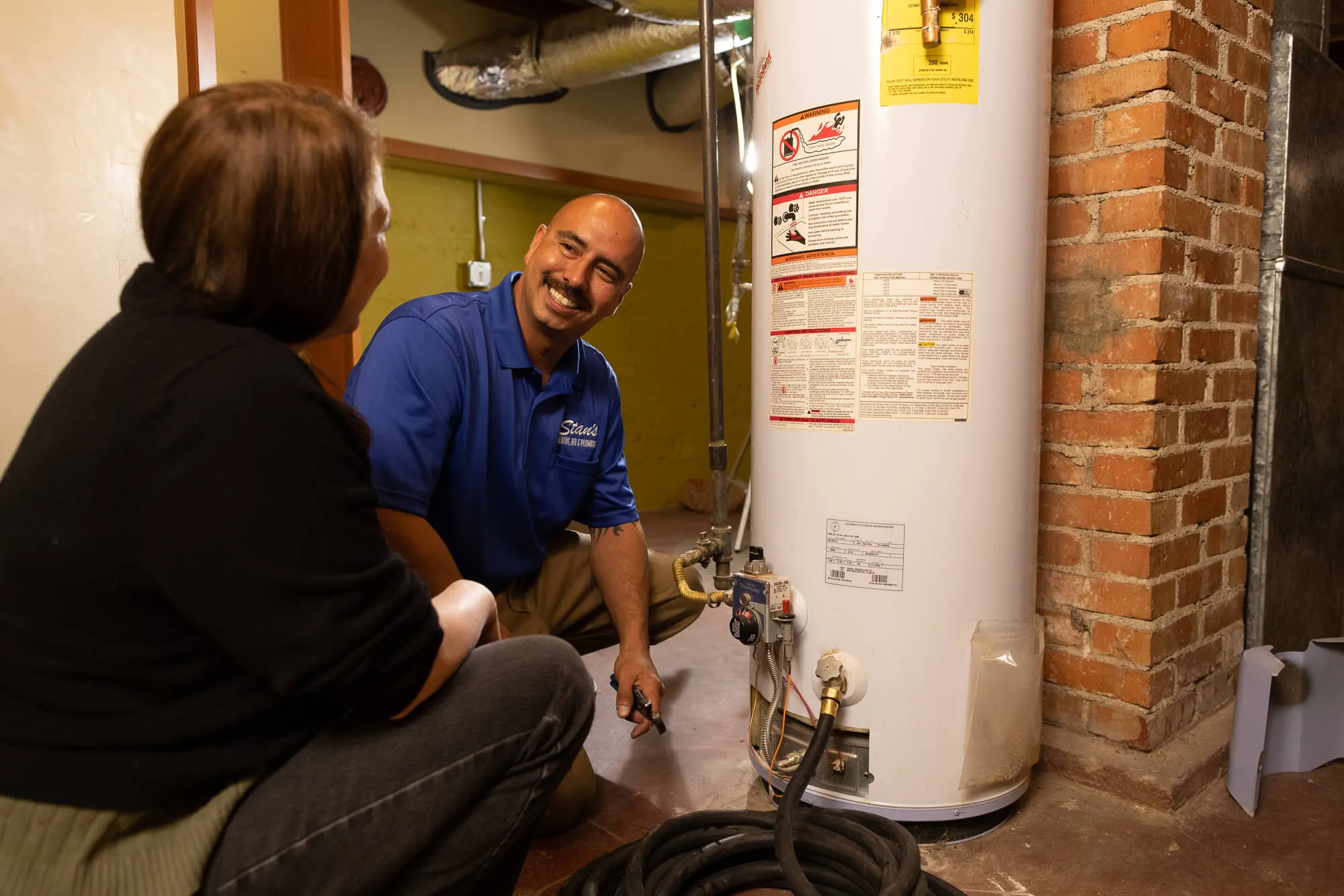 A technician in a blue shirt is inspecting a water heater while talking to a seated person in a basement.