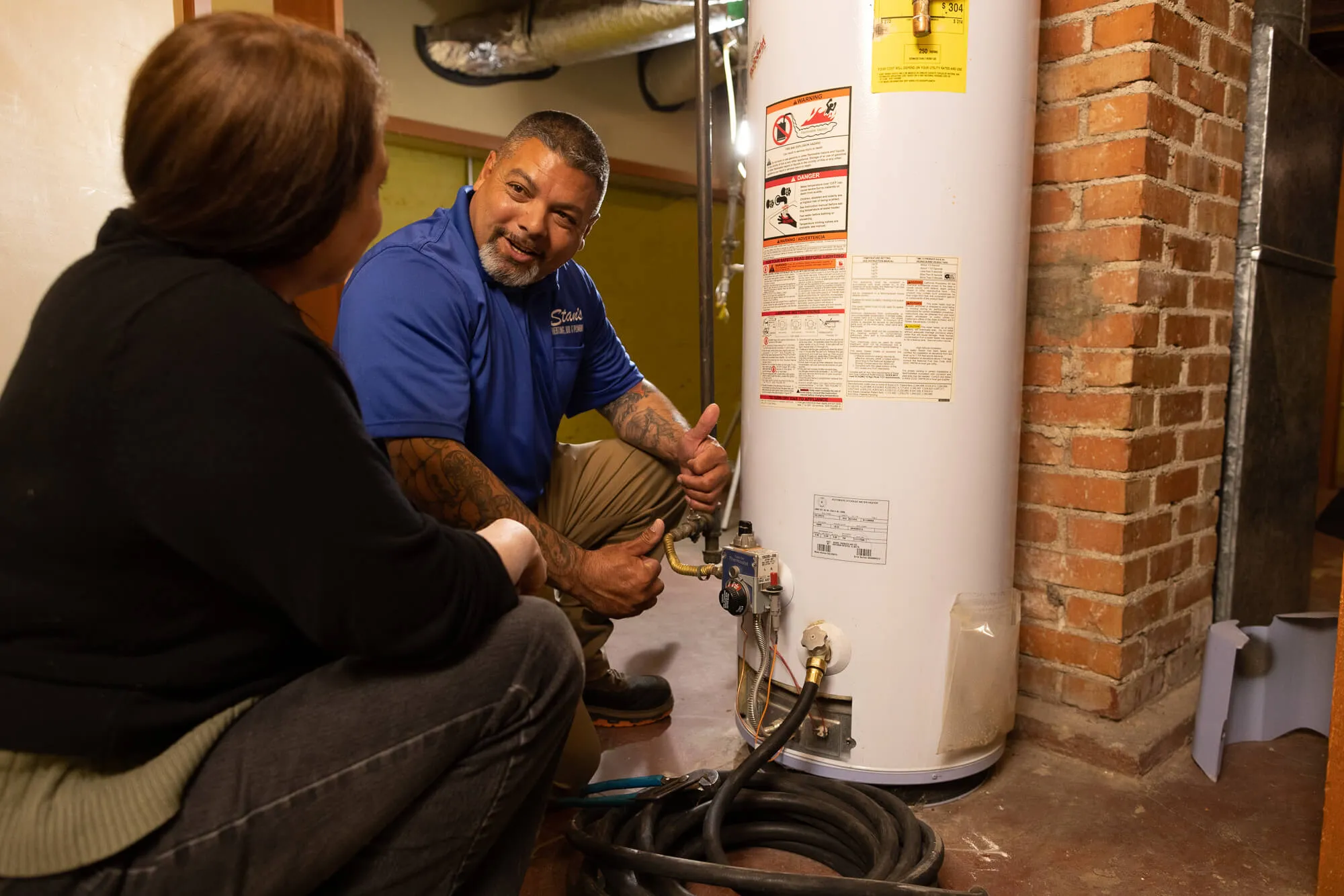 A plumber in a blue shirt explaining something about a water heater to a person sitting nearby.