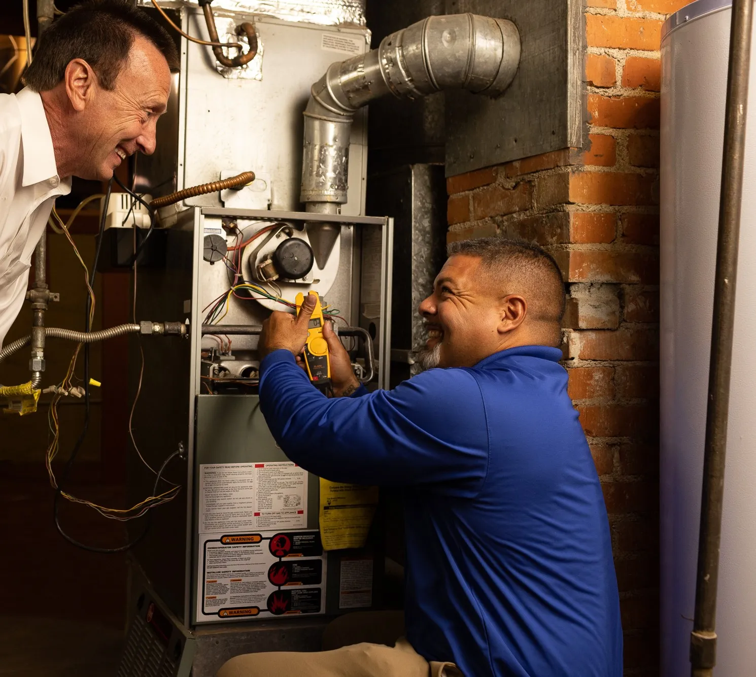 Two men working on a heating system in a basement, one holding a tool and the other observing.