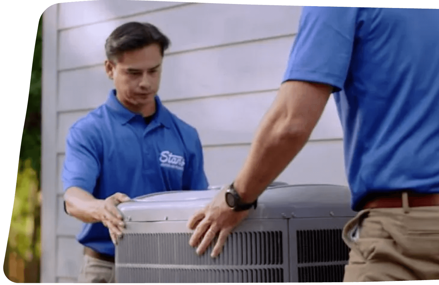 Two technicians in blue uniforms installing an outdoor air conditioning unit next to a house.