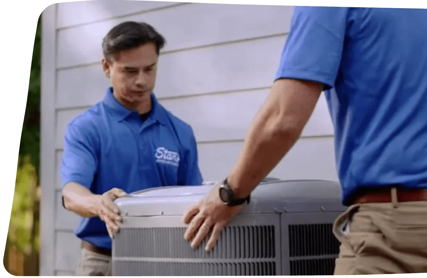 Two technicians in blue uniforms installing an outdoor air conditioning unit next to a house.