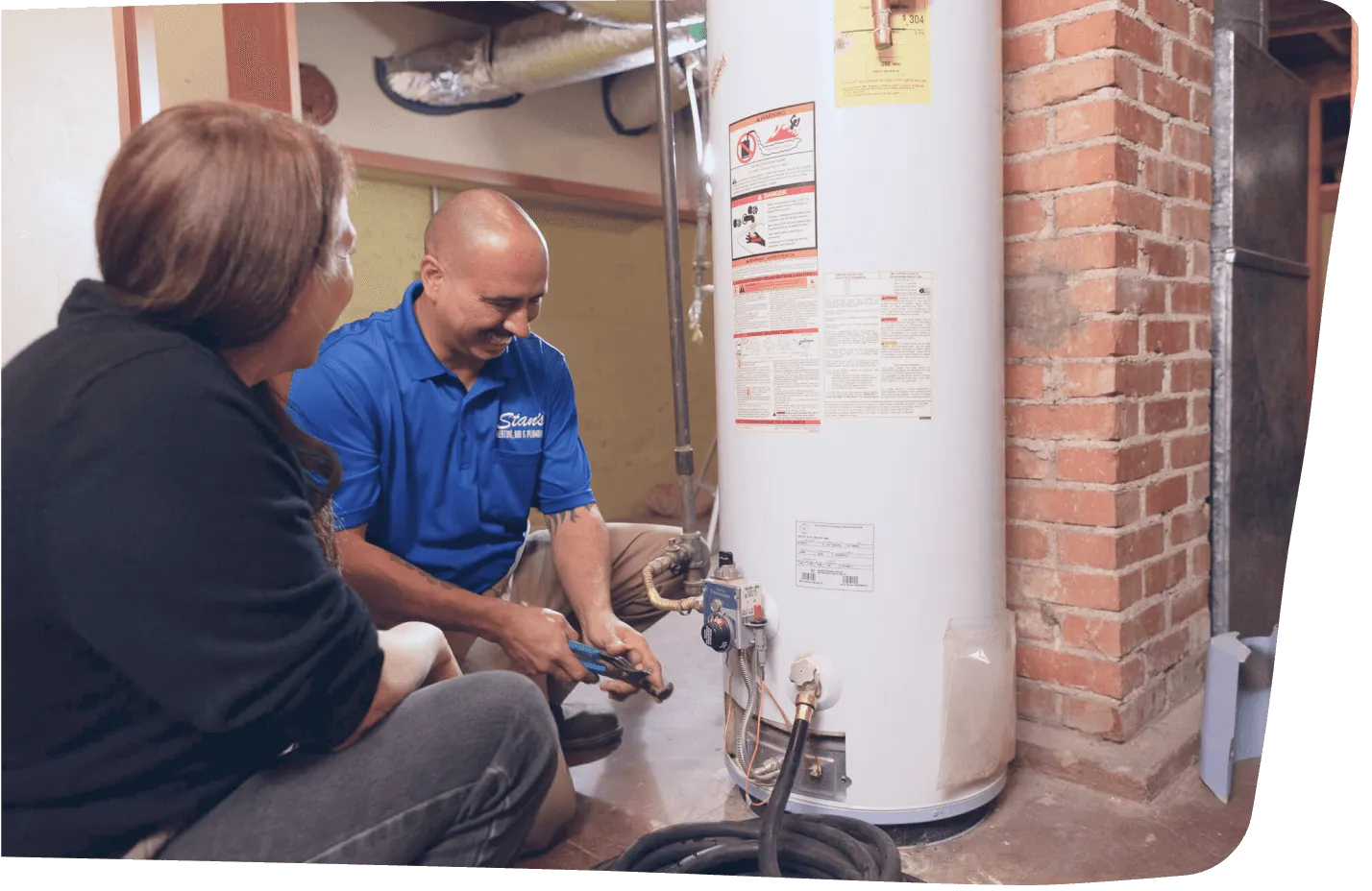Technician repairing water heater while explaining process to homeowner in basement setting.