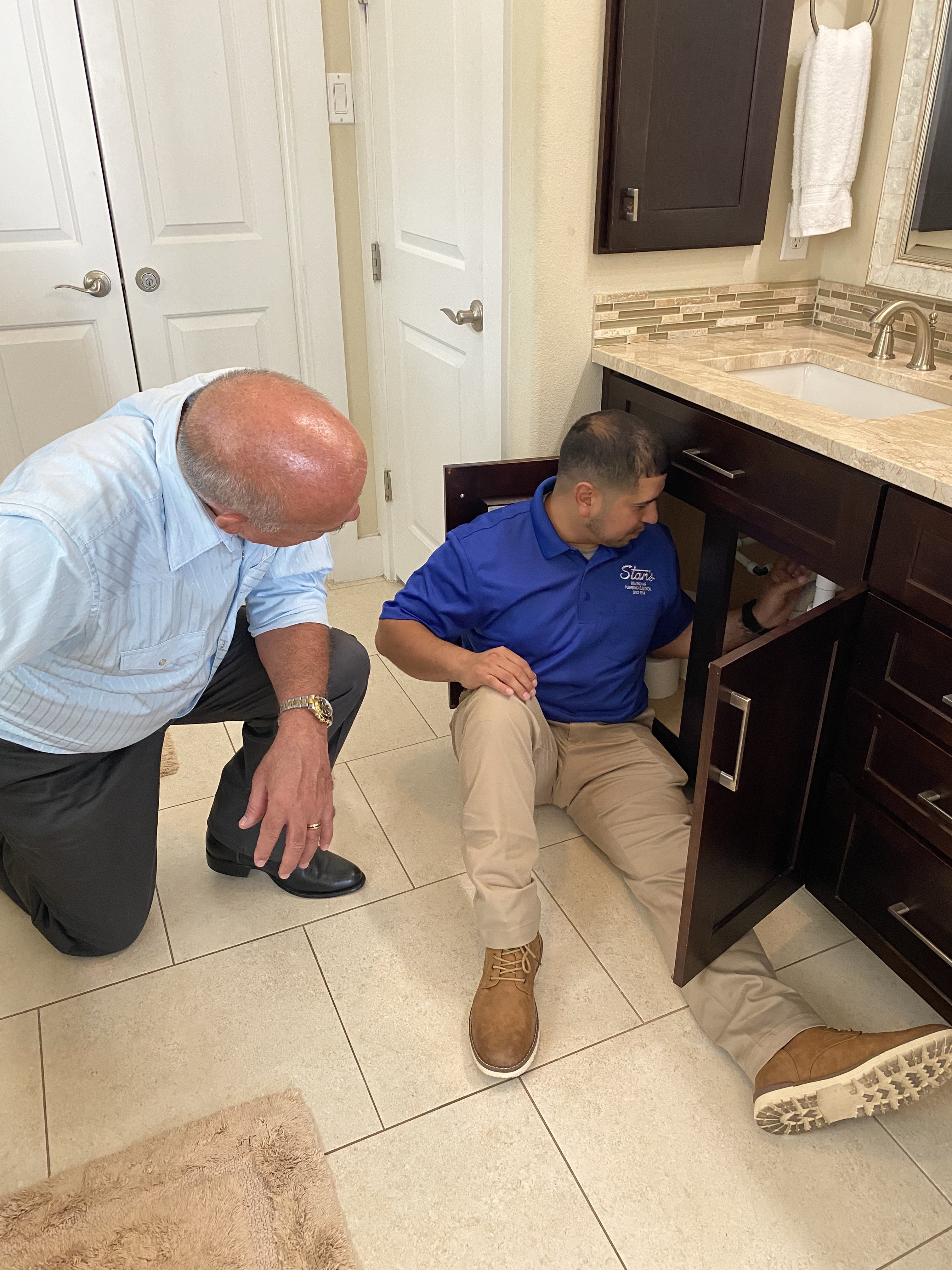 Plumber inspecting under-sink plumbing while homeowner observes in a modern bathroom setting.