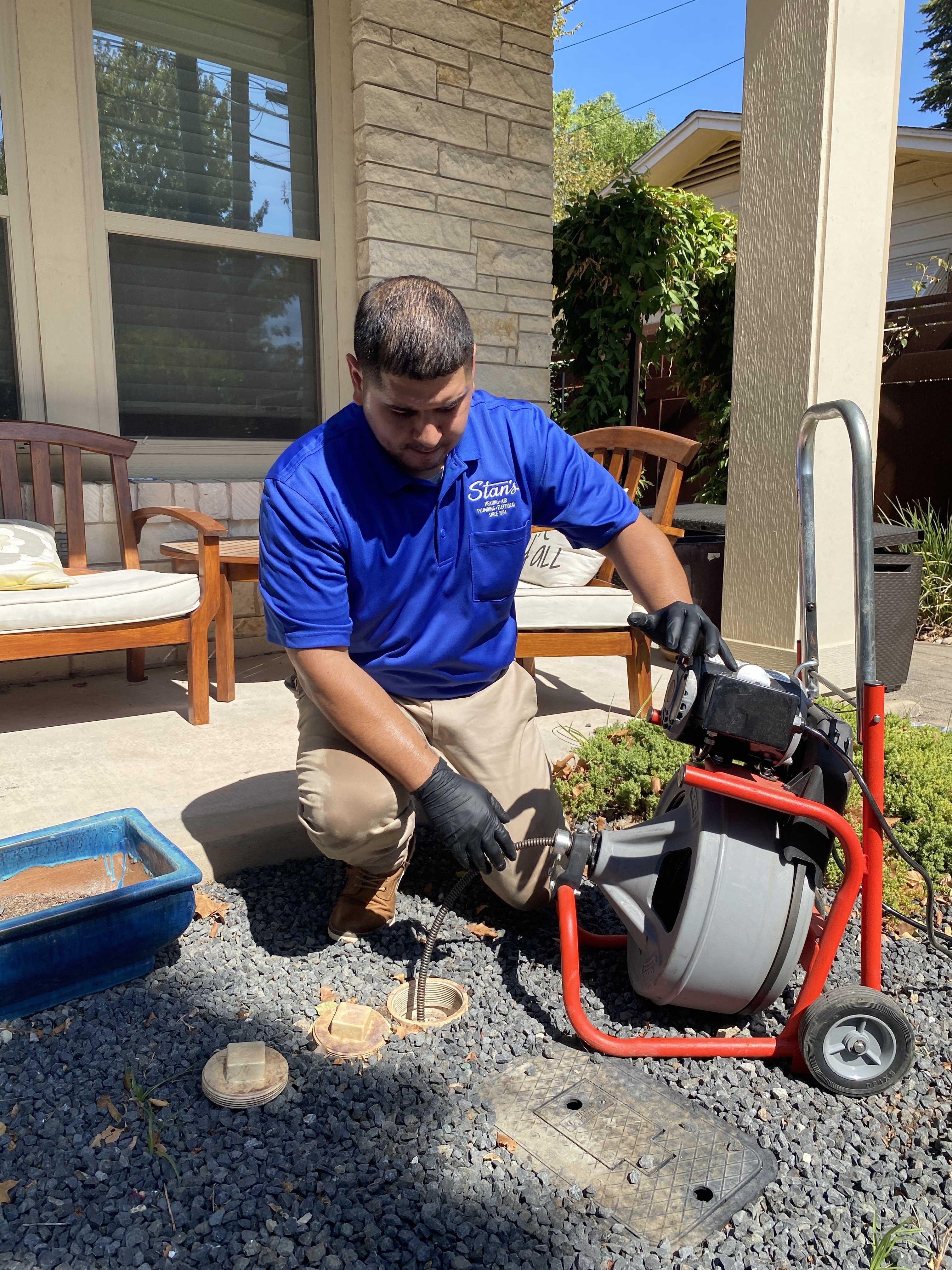Plumber in blue uniform using equipment to inspect outdoor drain near a house.
