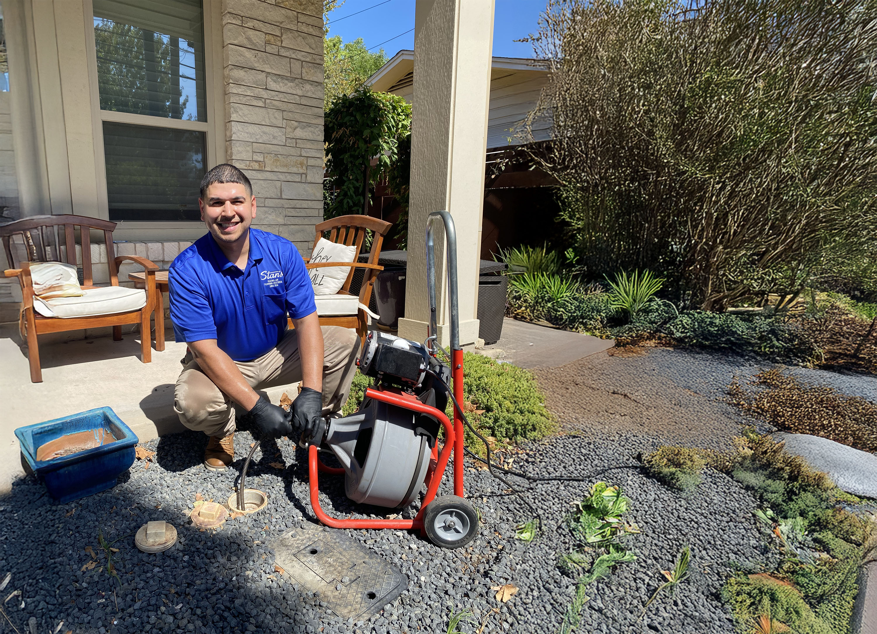 Technician clearing drain with plumbing snake outside a house on a sunny day.