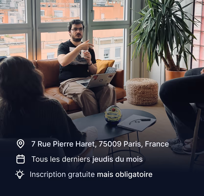 Personne discutant dans un salon avec un ordinateur portable sur les genoux, entourée de deux autres personnes et de grandes fenêtres donnant sur des bâtiments.