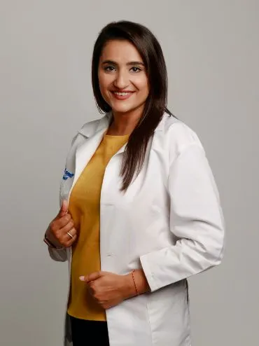 Dr. Shreya Khatri smiling in a white lab coat over a mustard yellow top against a plain gray background.
