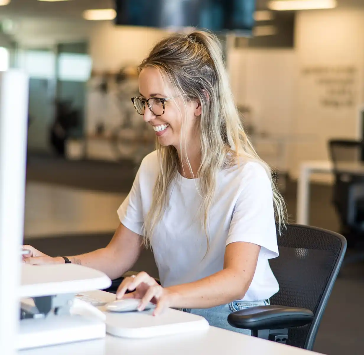 Smiling woman with glasses and blonde hair typing on a keyboard in a modern office.