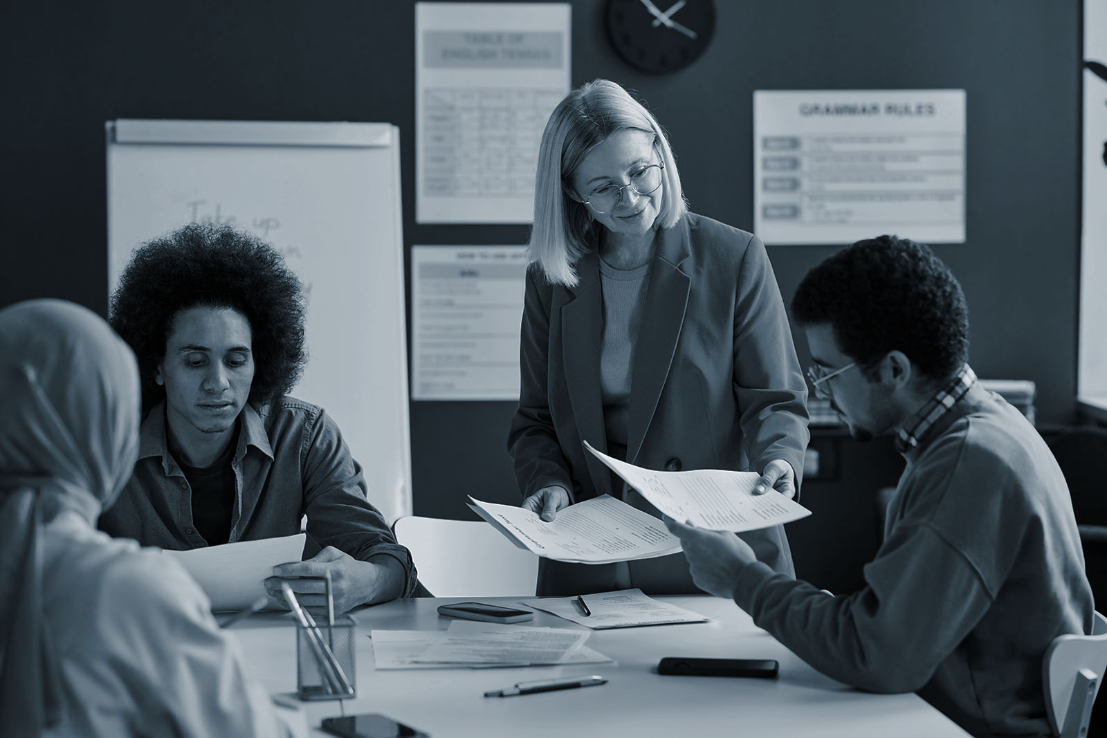 Mature teacher handing papers to students in a classroom, blue duotone