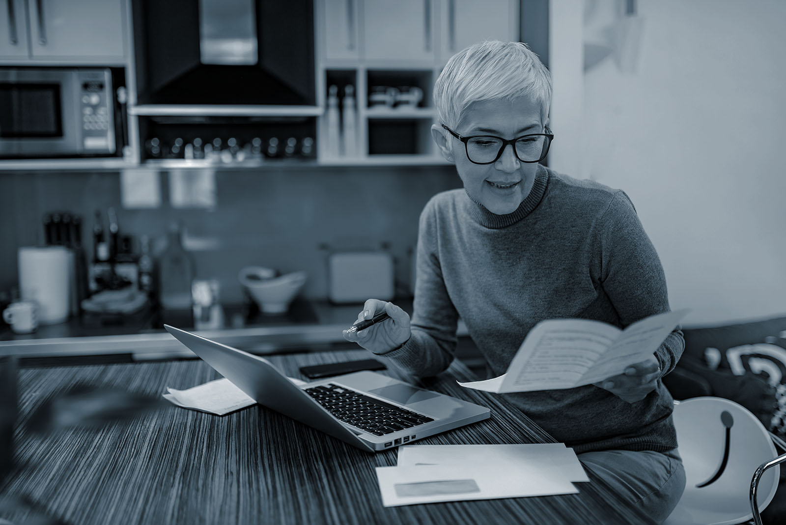 Senior woman at her kitchen table reviewing IRA beneficiary paperwork on a laptop, shown in Ignite's blue duotone style