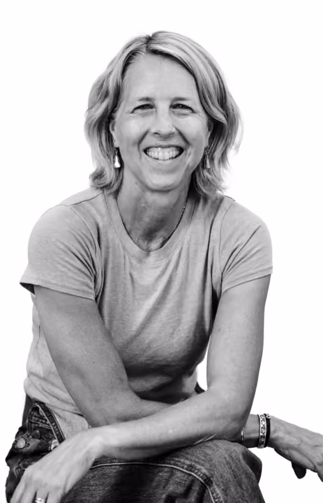 Smiling woman with short hair wearing a t-shirt and jeans, sitting with arms crossed over her knee.