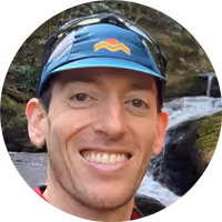 Smiling man wearing a blue cap with a colored logo, with a rocky stream in the background.
