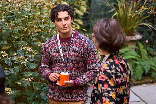 Man in patterned sweater holding orange cup talks to woman in floral jacket outdoors near plants.