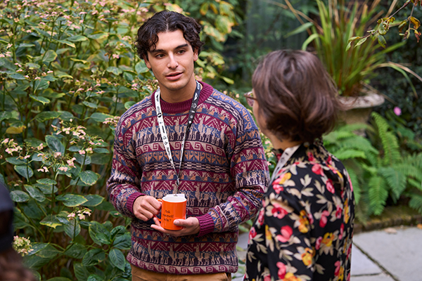 Man in patterned sweater holding orange cup talks to woman in floral jacket outdoors near plants.