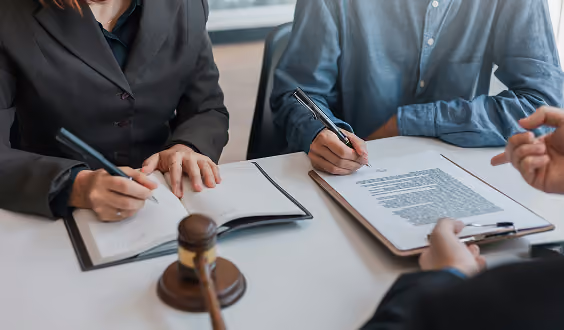 Three people signing legal documents at a table with a judge's gavel in the foreground.