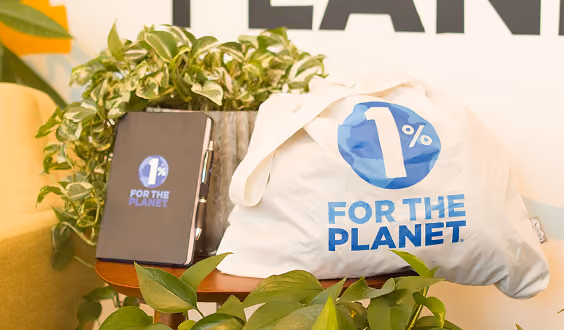 White bag and black notebook with blue 1% For The Planet logo on a wooden table with green plants.