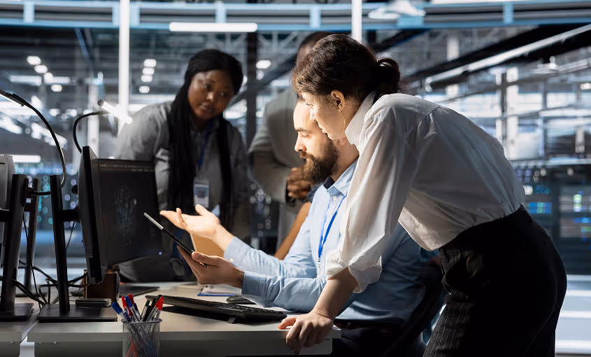 Three colleagues in an office collaborating around a computer monitor, with one woman leaning over the desk holding a tablet.