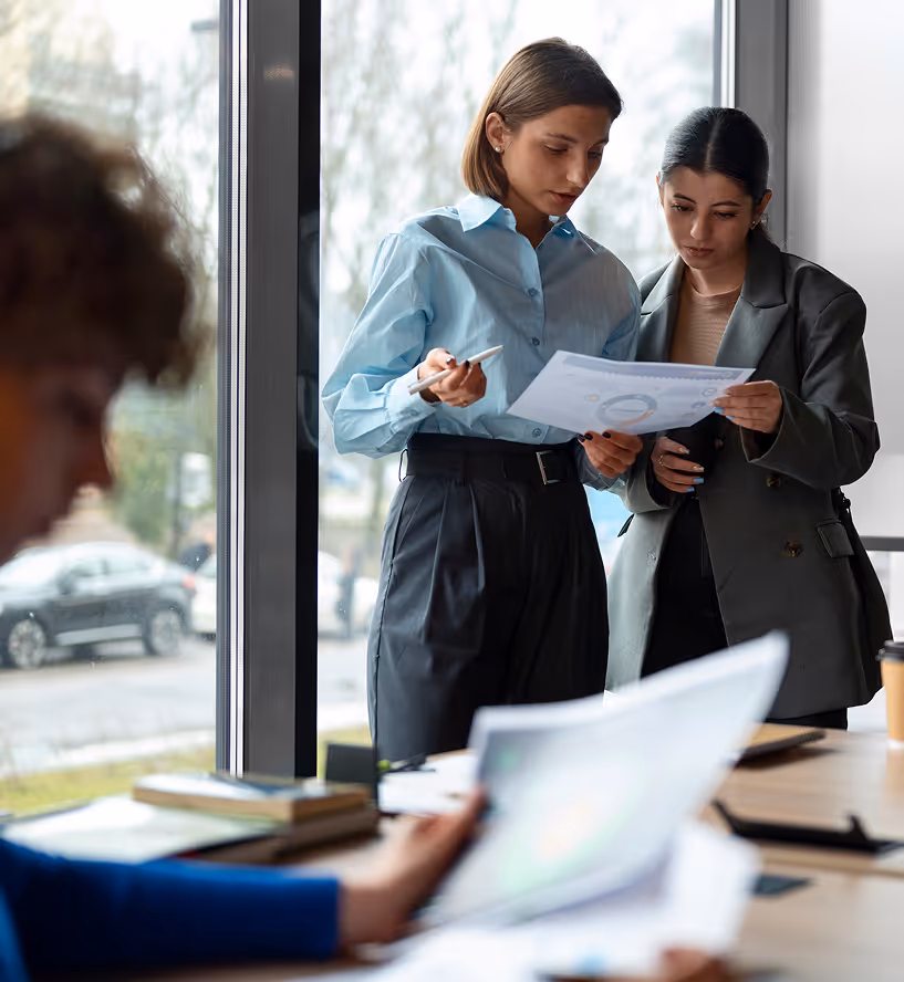 Two women standing by a window examining a document together in an office setting.
