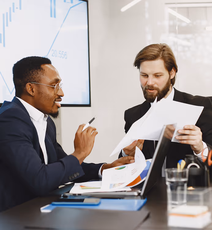 Two businessmen discussing charts and documents over a laptop in a modern office.