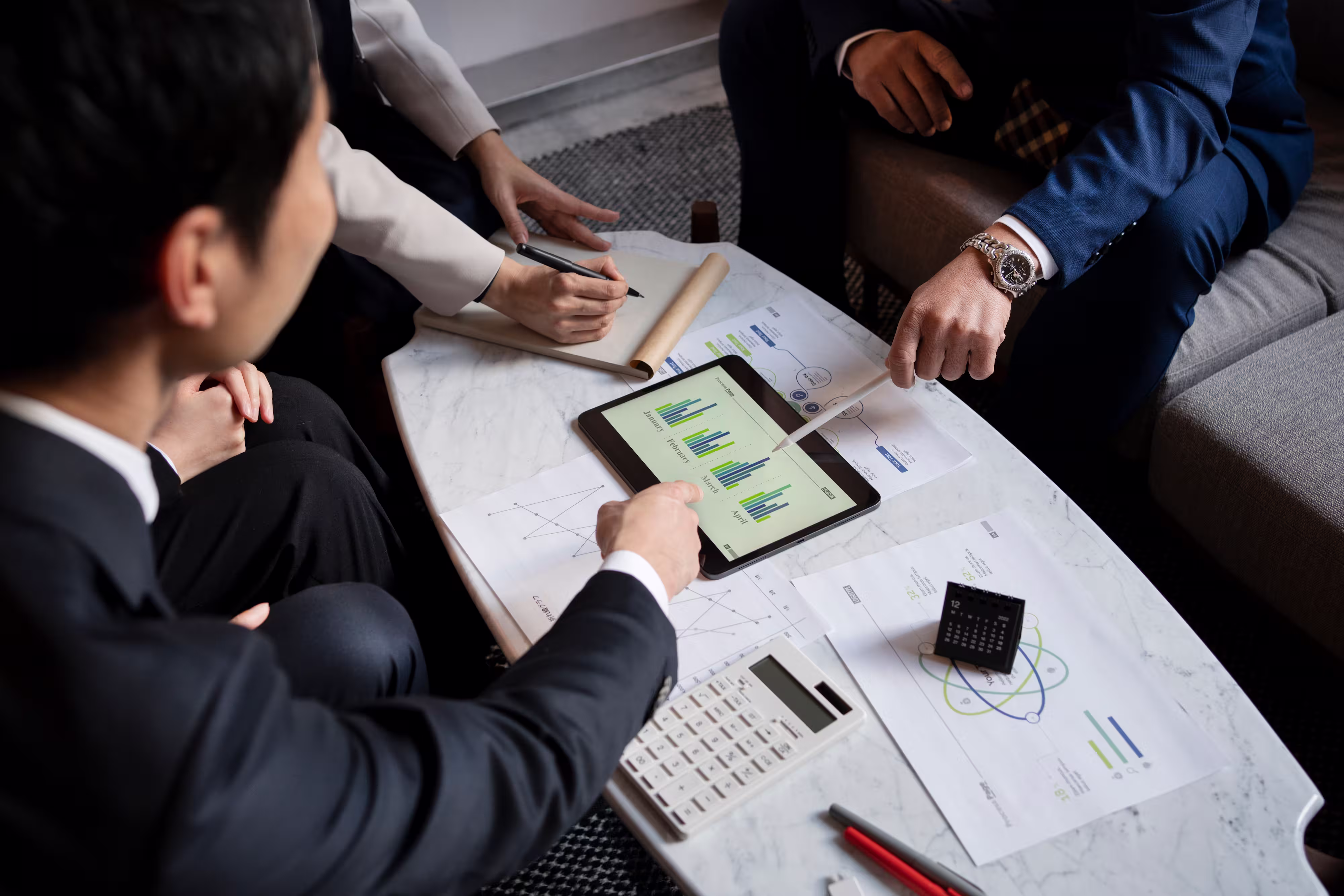 Group of business professionals discussing charts and data on a tablet and documents around a white table.