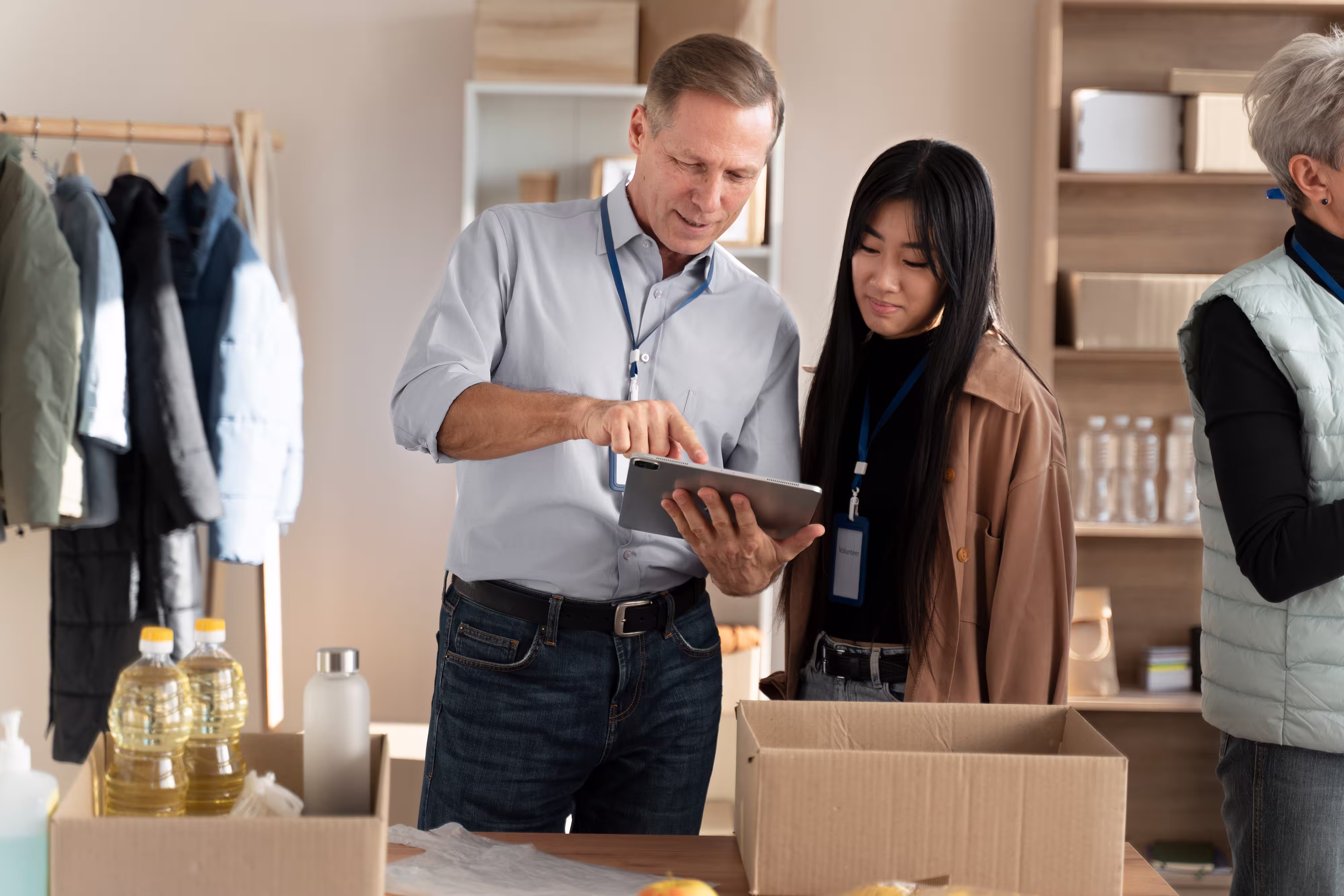 Two volunteers looking at a tablet while packing food items into cardboard boxes in a room with shelves and coats hanging.