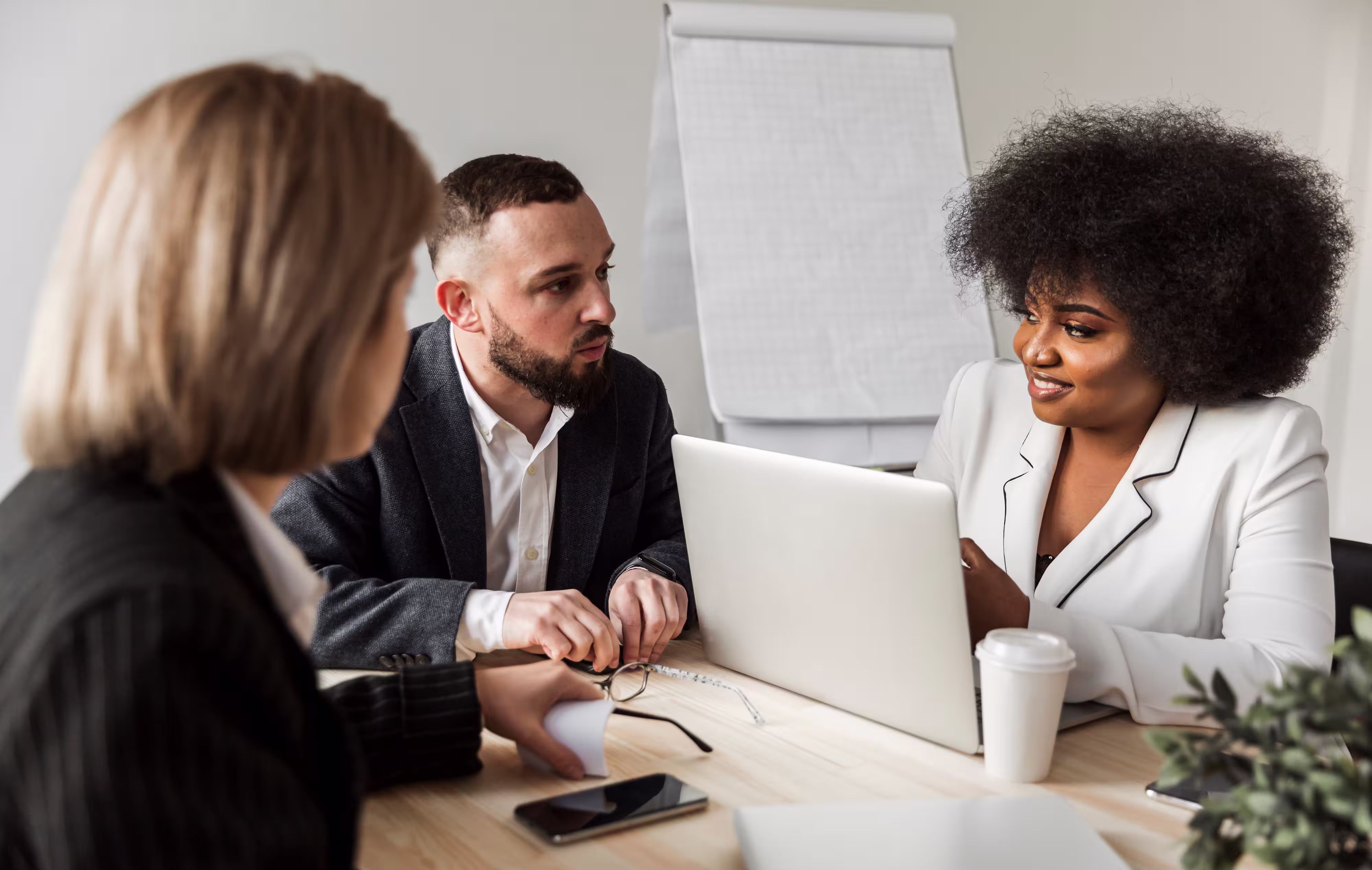 Three professionals engaged in a discussion around a table with a laptop and coffee cup.