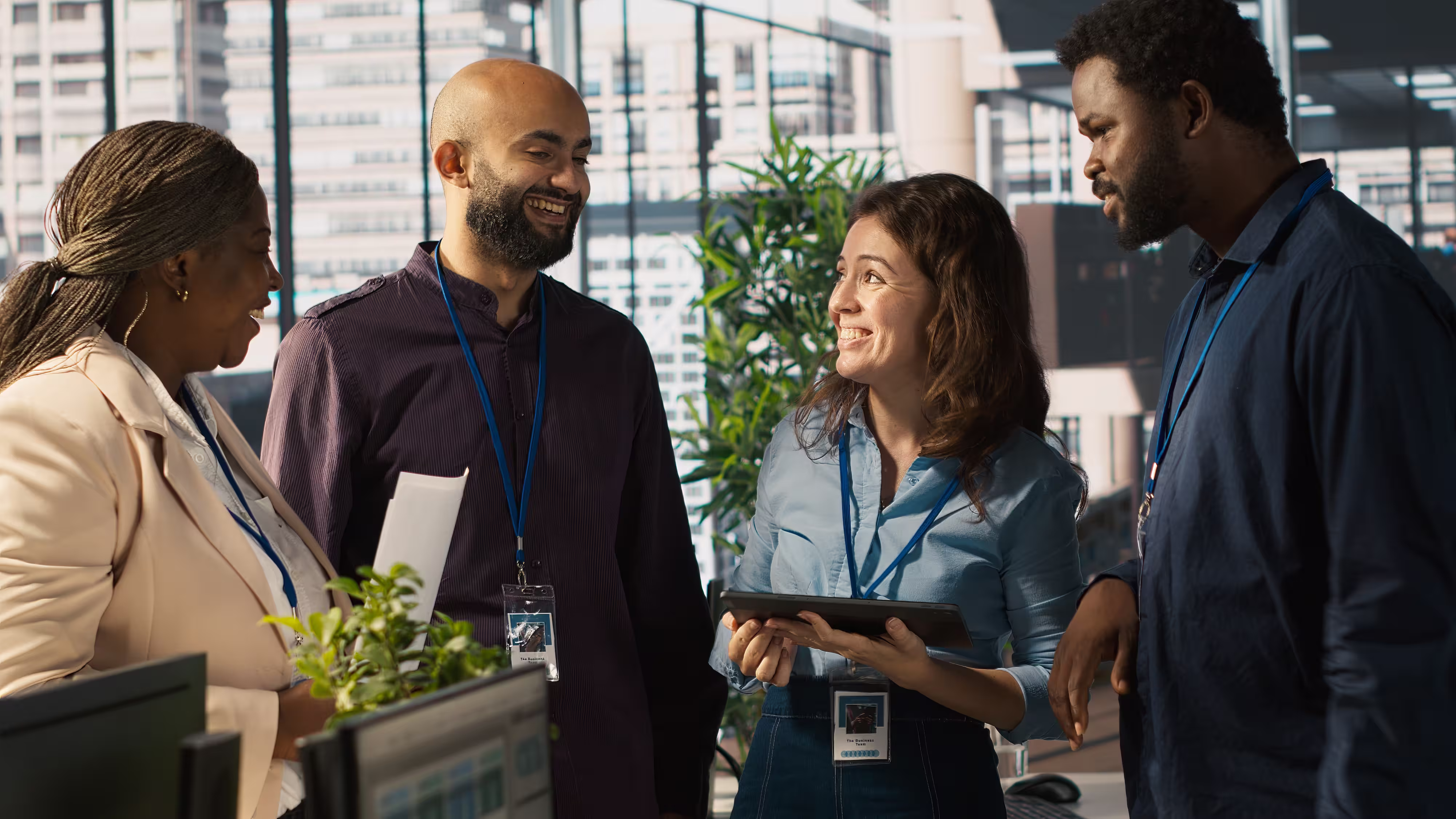 Four diverse coworkers smiling and talking in an office with a tablet and documents in hand.