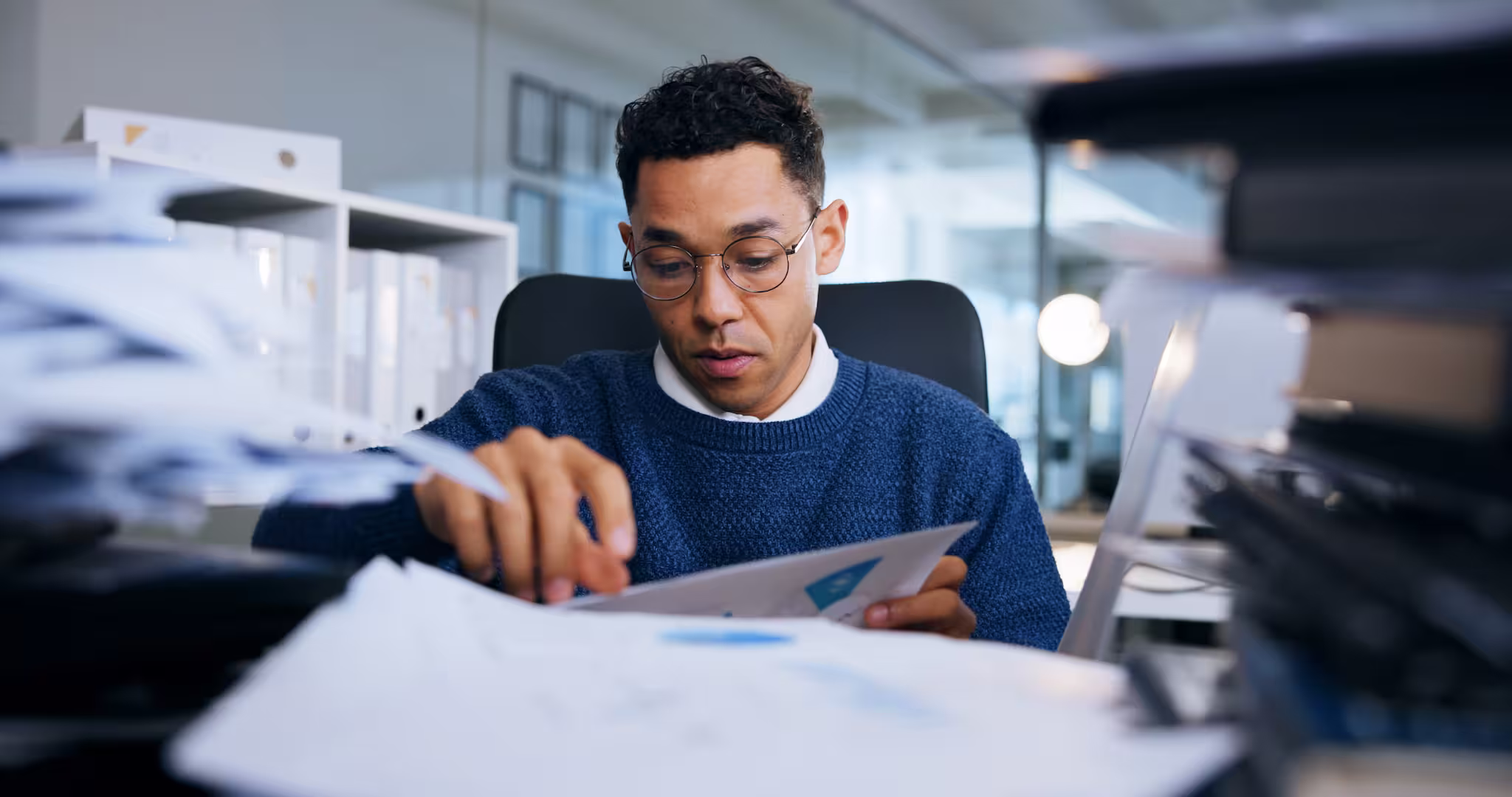 Man with glasses in a blue sweater reviewing documents at a cluttered office desk.