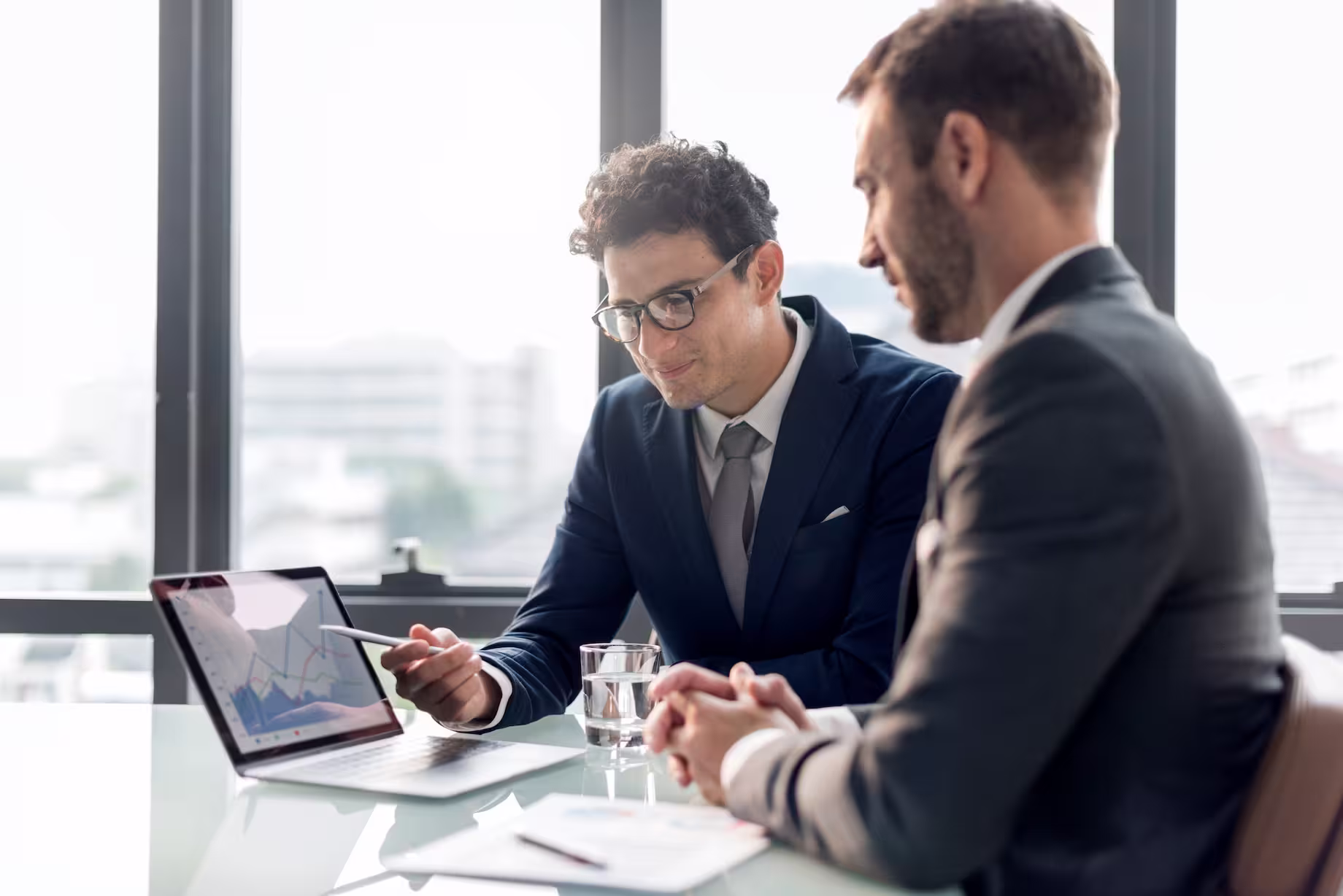 Two businessmen in suits reviewing data charts on a laptop in a bright office.