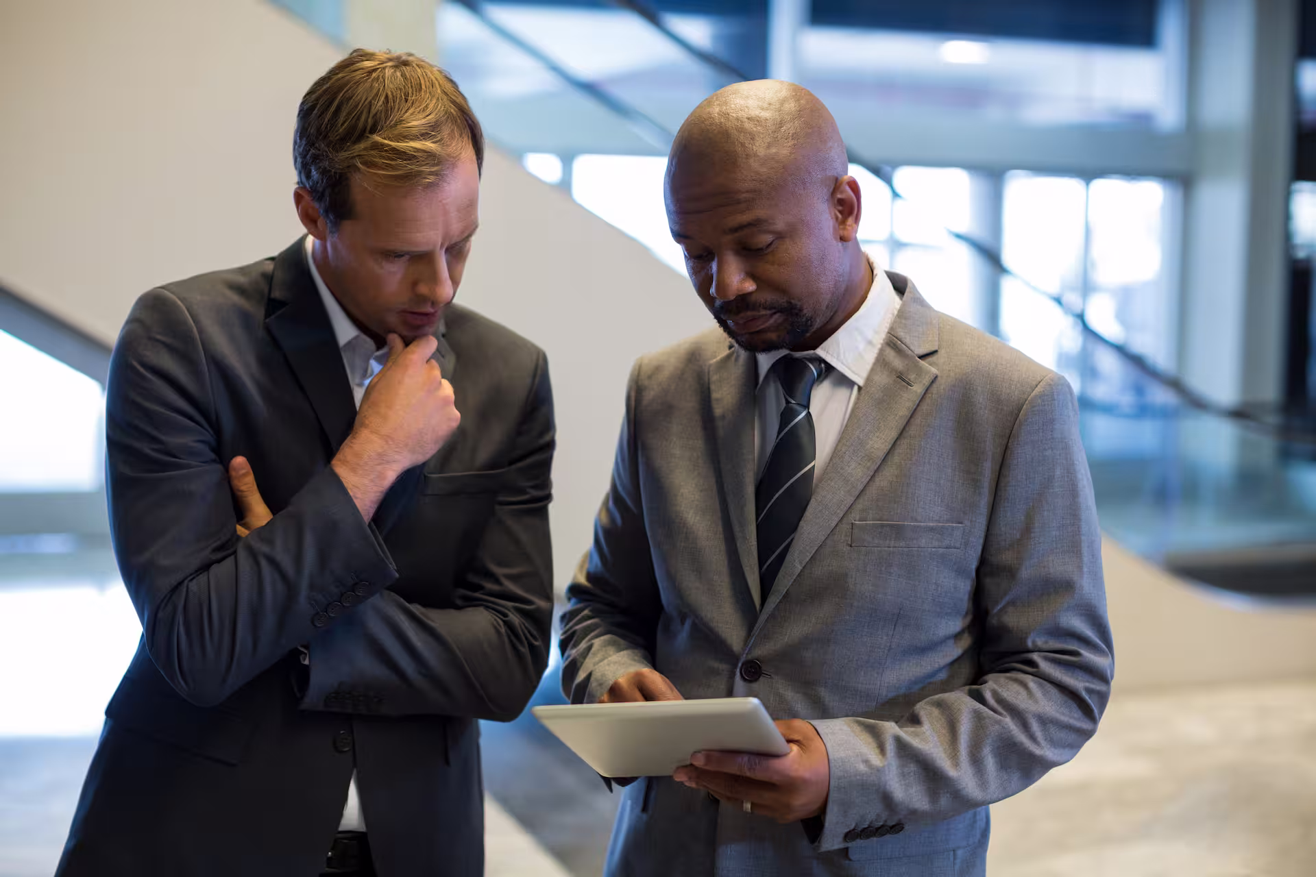 Two businessmen in suits looking at a tablet in a modern office setting.