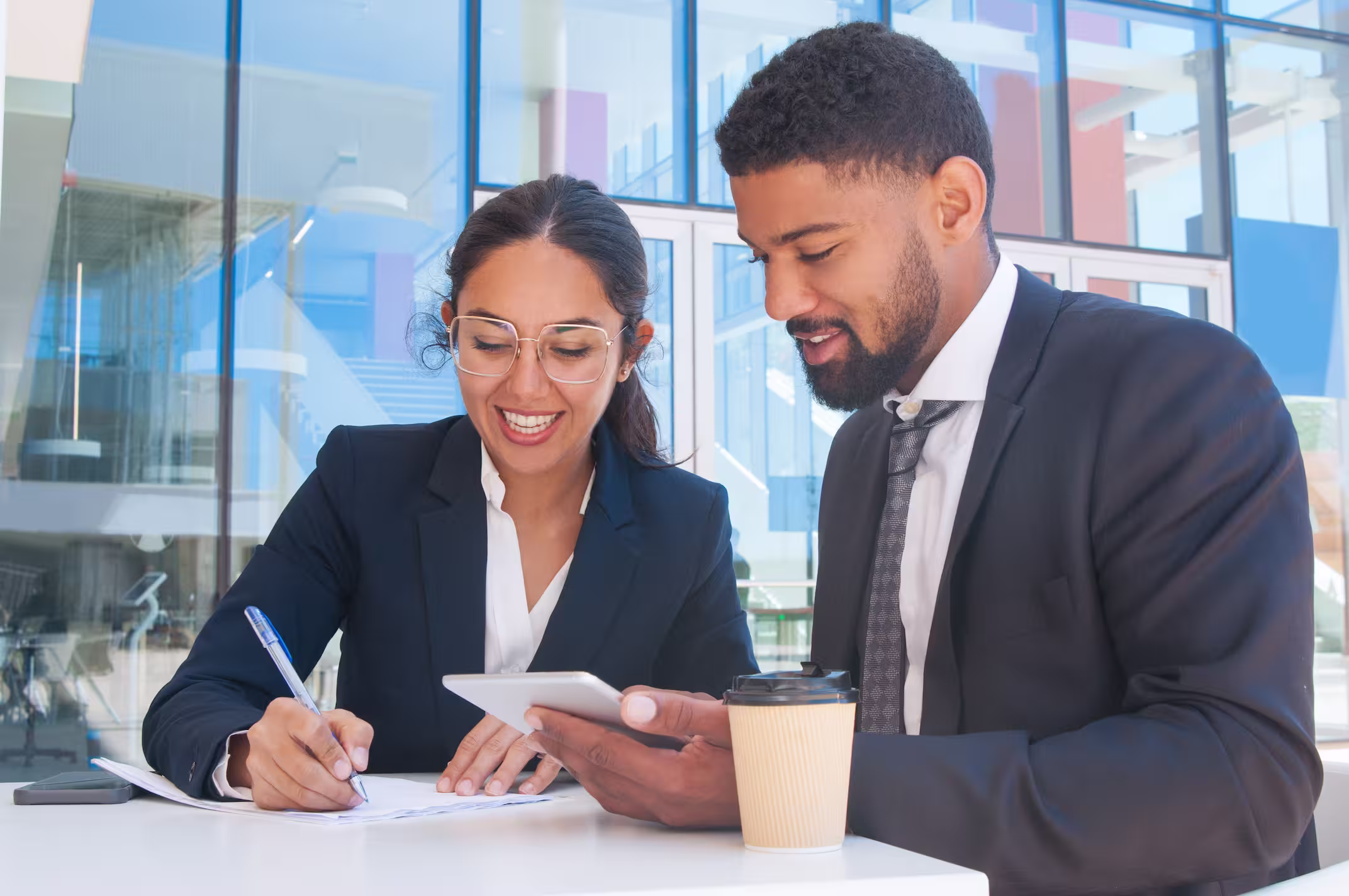 Two business professionals in suits collaborating at a table with a tablet and documents in a modern office setting.