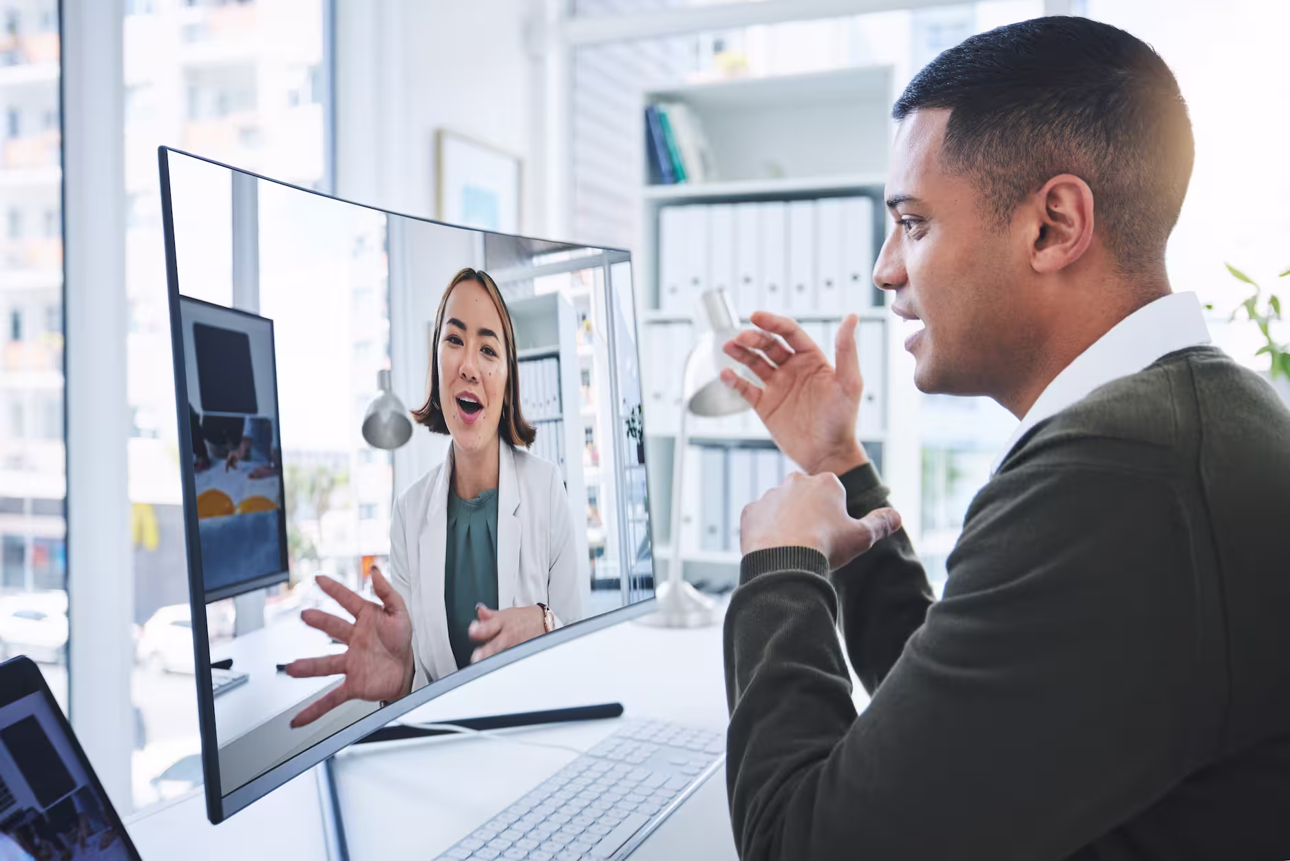 Man in an office having a video call with a woman displayed on a curved monitor.