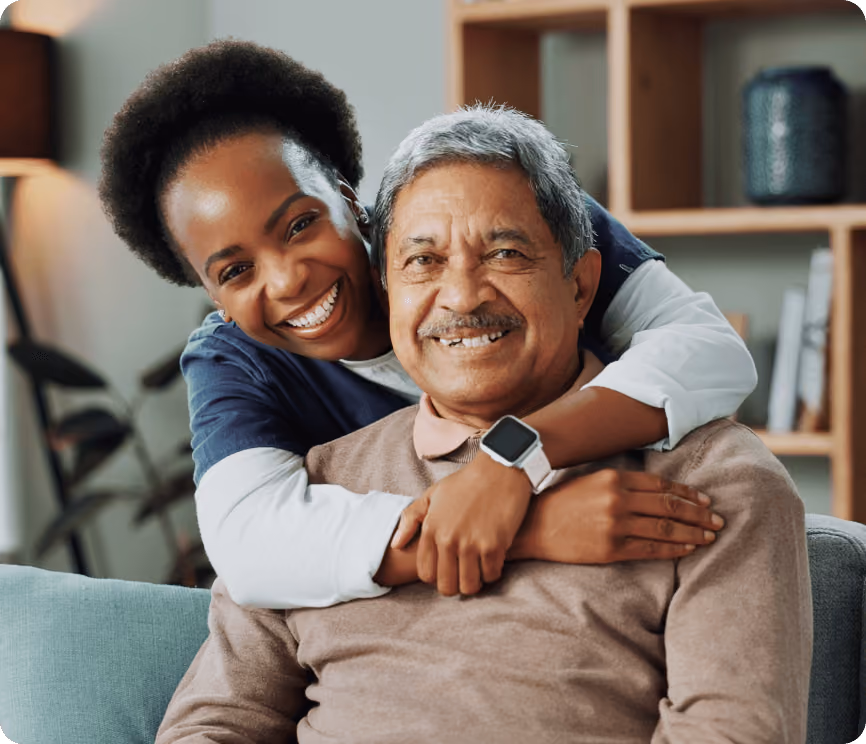 A caregiver hugging her patient from behind. They are both smiling at the camera.