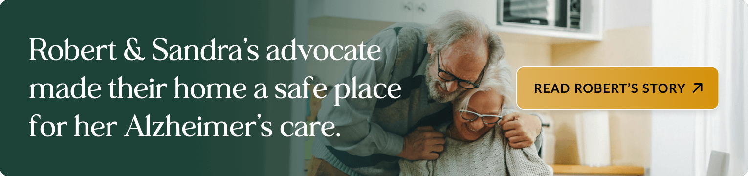 Elderly couple embracing in a bright kitchen, with the man wearing glasses and a blue cardigan standing behind the woman who is wearing glasses and a cream sweater. Banner text: Robert & Sandra's advocate made their home a safe place for her Alzheimer's care. Includes a button: READ ROBERT'S STORY.
