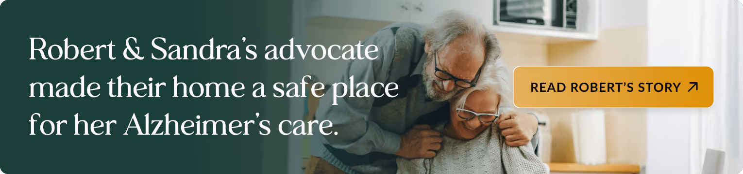 Elderly couple embracing in a bright kitchen, with the man wearing glasses and a blue cardigan standing behind the woman who is wearing glasses and a cream sweater. Banner text: Robert & Sandra's advocate made their home a safe place for her Alzheimer's care. Includes a button: READ ROBERT'S STORY.
