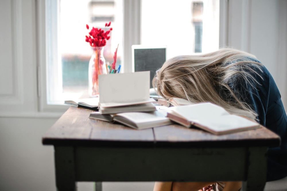 An exhausted woman, face down on a table loaded with books and papers.