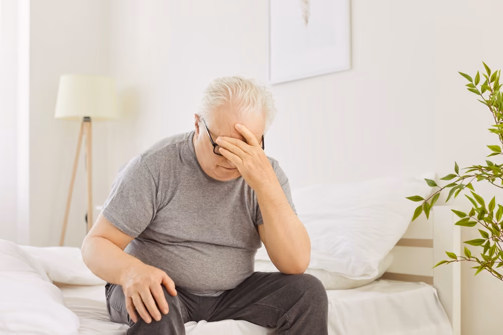 Older white man sitting on the edge of his bed, tired