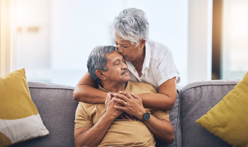 A senior man on the couch being embraced by his partner from behind.
