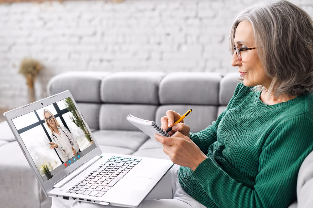 Older woman taking notes during a video appointment with a physician on a laptop while seated on a couch at home.