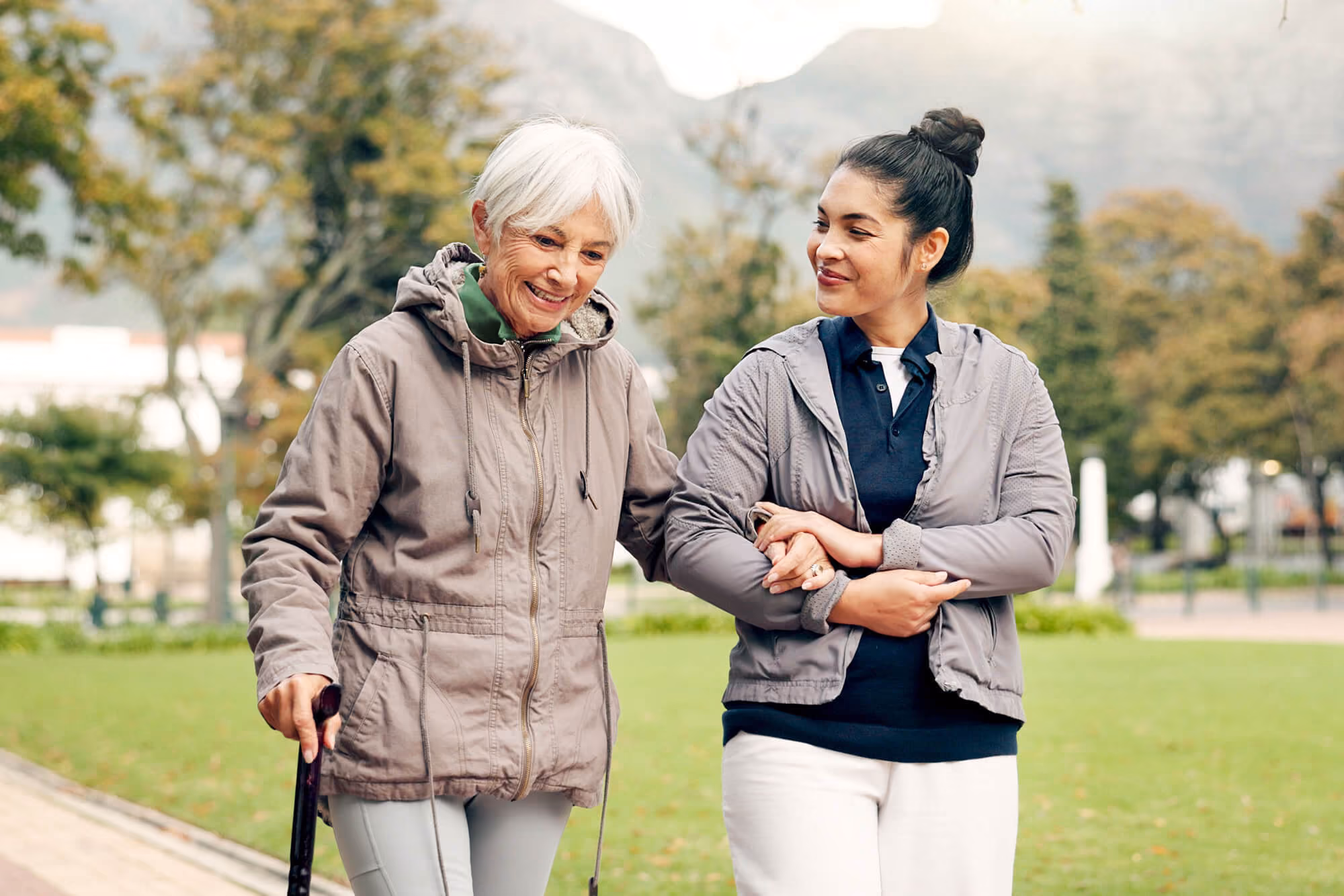 Older woman using a cane walks outdoors alongside a younger woman offering support, both smiling in a park setting.