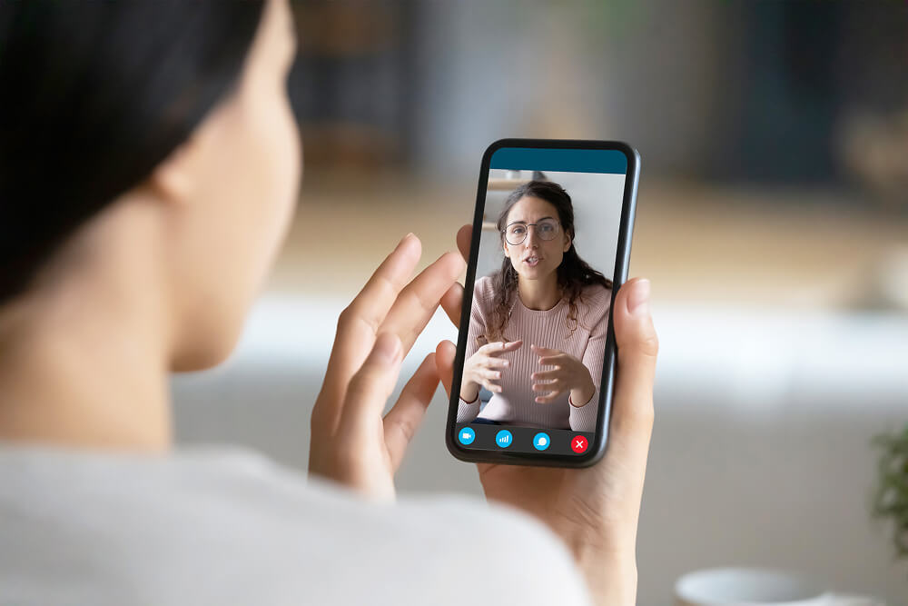 Person holding a smartphone during a video call with a woman wearing glasses who appears to be explaining something, seen speaking and gesturing on the screen.